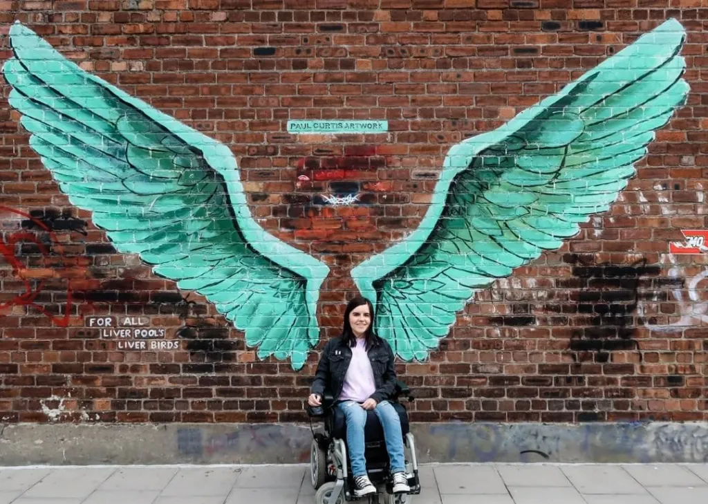 photo of blogger Emma Muldoon in a wheelchair wearing a pink shirt black jacket and blue jeans in front of a mural painted on a brick wall that makes her look like she has large green wings