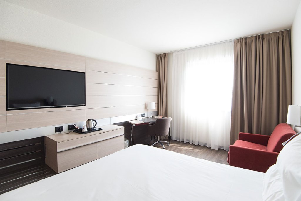 picture of a hotel room with bed desk flat screen tv beige curtains and a red chair
