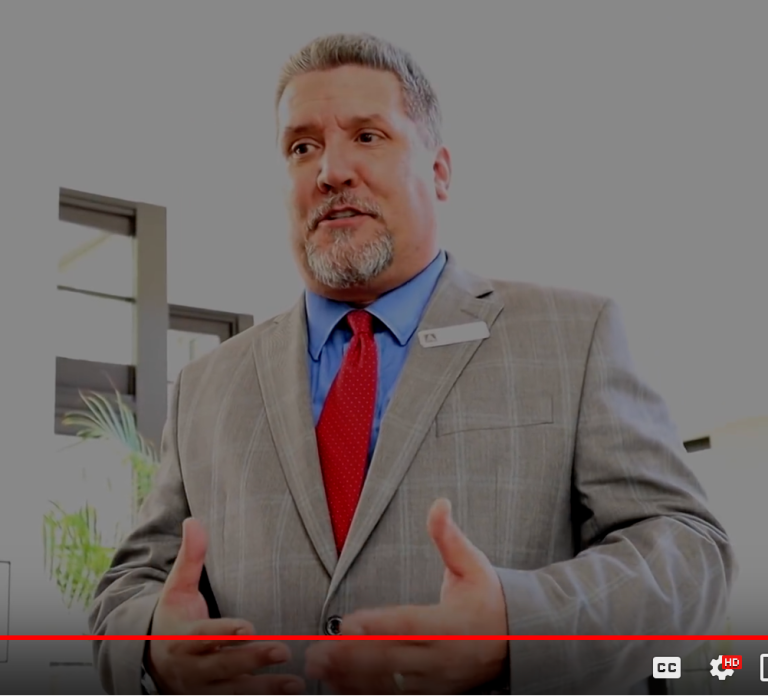 screen grab from YouTube video of Jim Alfond, Hotel Manager. He is wearing a tan suit, blue shirt and red tie. His hair is graying and he has a goatee